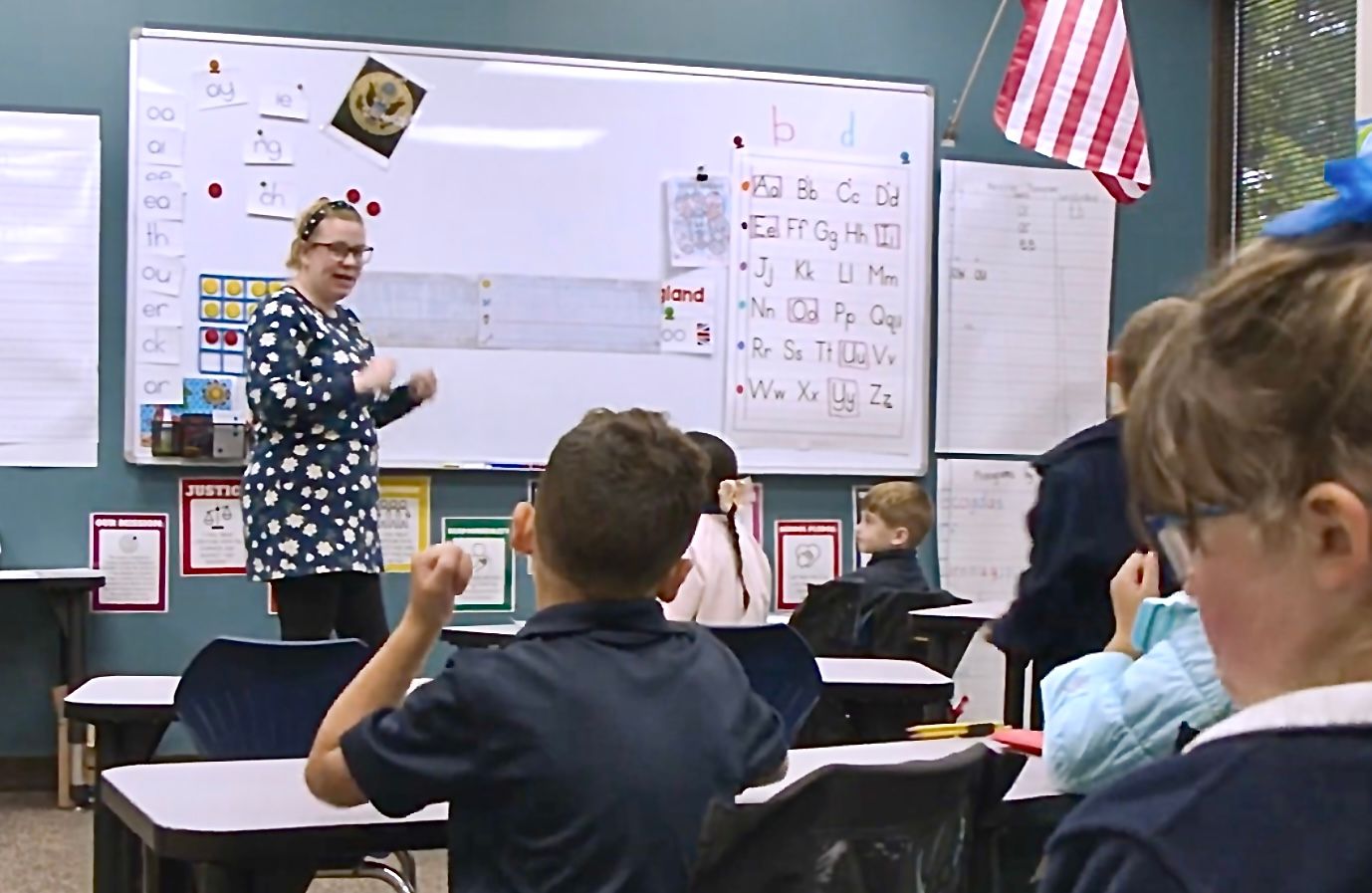 An ACAR teacher leading a lesson from the front of the room, students at their desks with a hand raised, phonogram cards and the printed and cursive alphabets on the board behind her
