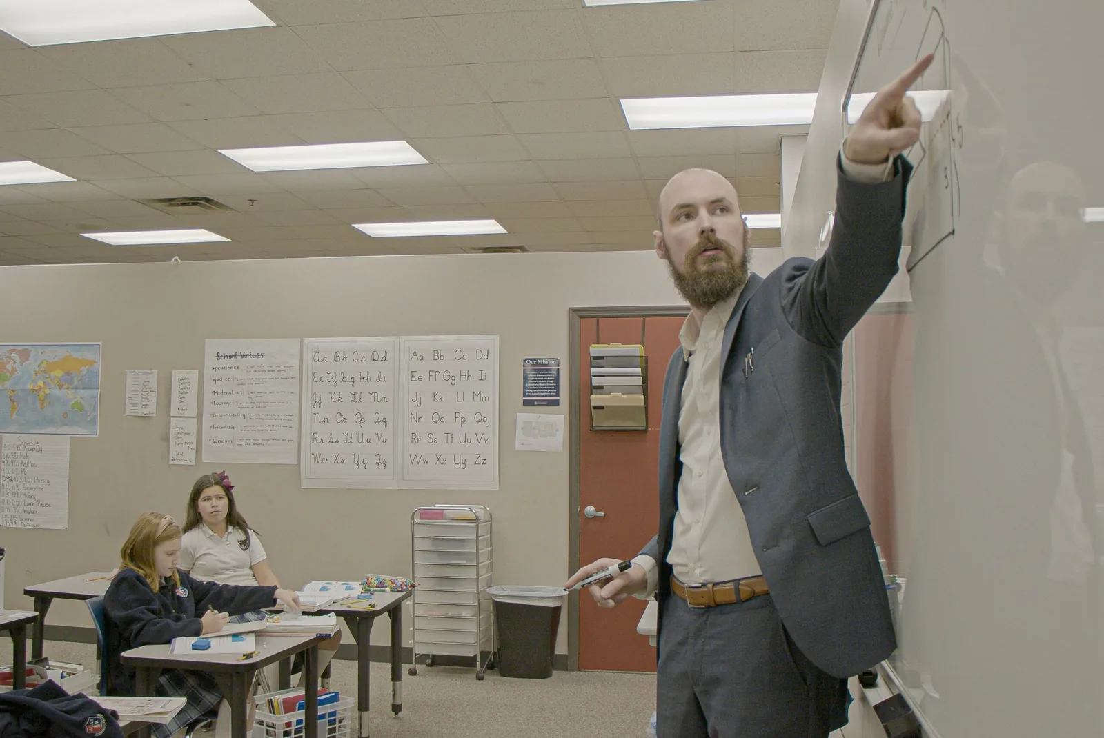 An ACAR teacher at the whiteboard, with the school virtues, cursive alphabet charts and a world map on the classroom wall