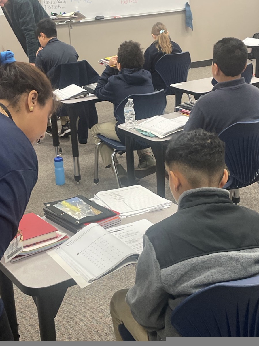 An ACAR teacher leaning over a desk to help a student with a workbook while the rest of the class works